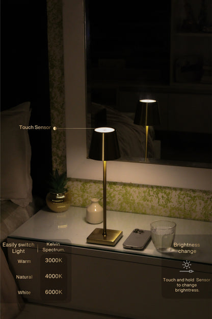 A modern table lamp with a black conical shade, gold-toned metal stem, and square base, shown lit on a bedside table in a dimly lit bedroom. The image includes callout text and overlays explaining touch sensor, Kelvin spectrum light, brightness change, and easy switch light features.