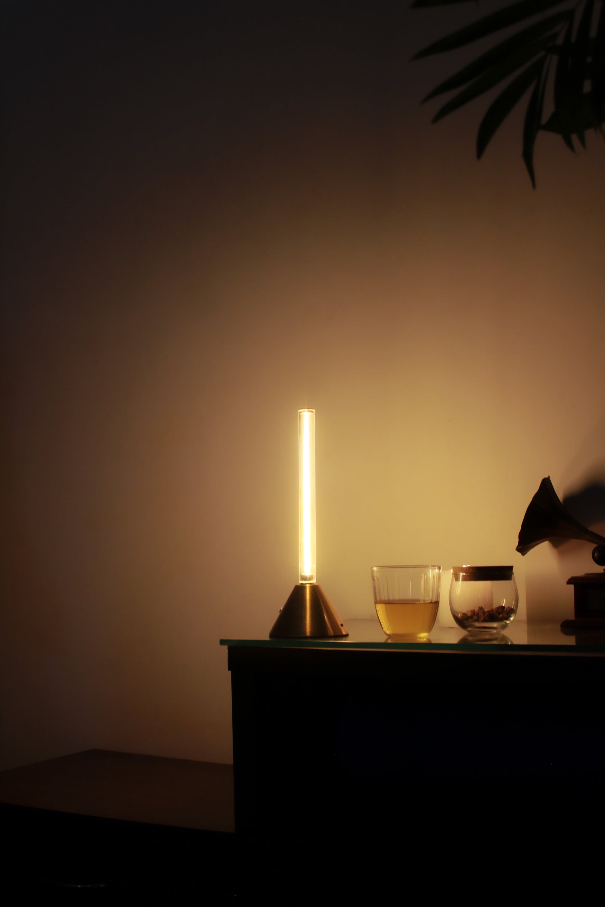 Metal Touch Modern table lamp with a warm glow on a dark surface, with a glass and a plant in the background.